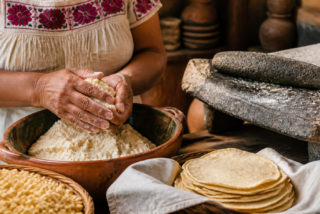 Manos muelen maíz nixtamalizado en un metate junto a tortillas recién hechas.