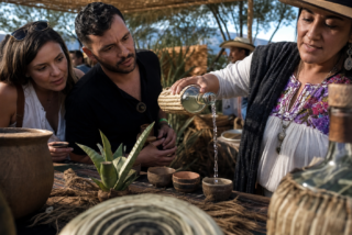 Productor vierte mezcal en una copita mientras visitantes observan la degustación en un festival al aire libre con agaves y artesanía regional.