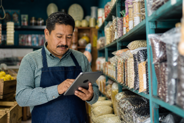 Pequeña tienda con anaqueles etiquetados y una persona registrando existencias en una tableta.