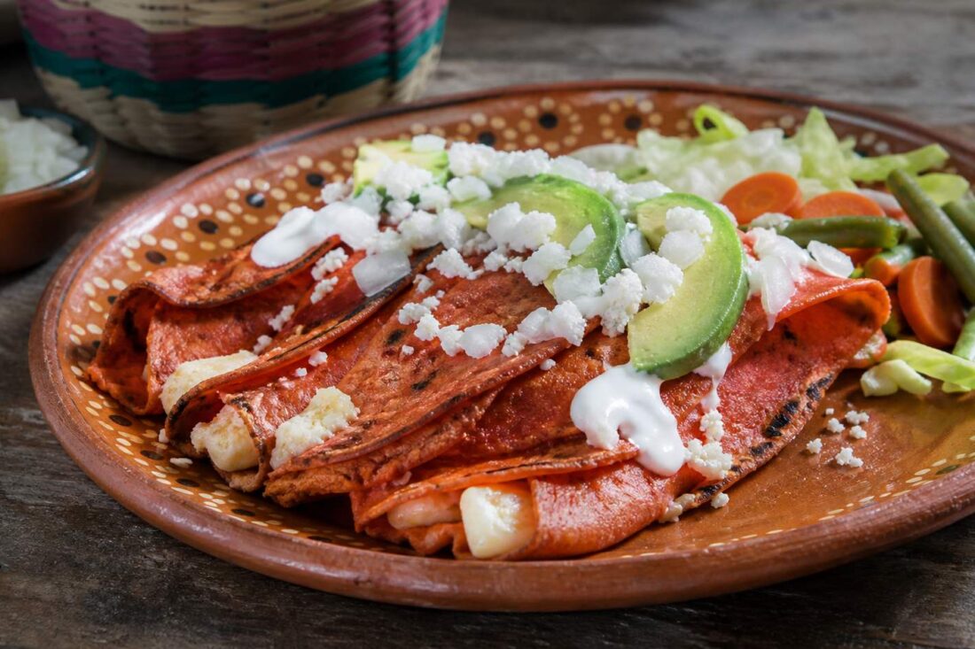Enchiladas potosinas de masa rojiza con chile, queso, crema, aguacate y vegetales en plato de barro