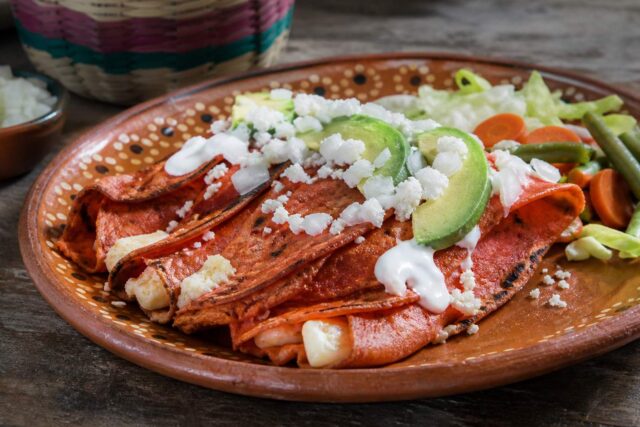 Enchiladas potosinas de masa rojiza con chile, queso, crema, aguacate y vegetales en plato de barro