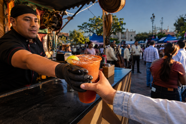 Vaso de michelada escarchado en una plaza de Villagrán, con puestos de comida, música y visitantes en una escena documental mexicana.
