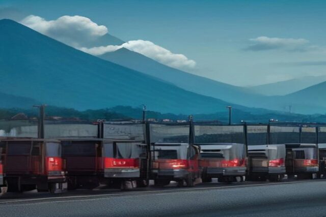Fila de tractocamiones rojos detenidos sobre una carretera con montañas y cielo azul con nubes al fondo