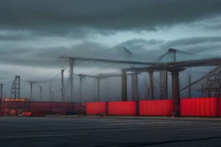 Patio industrial portuario con grúas, estructuras metálicas y contenedores rojos bajo un cielo nublado, sin personas ni texto