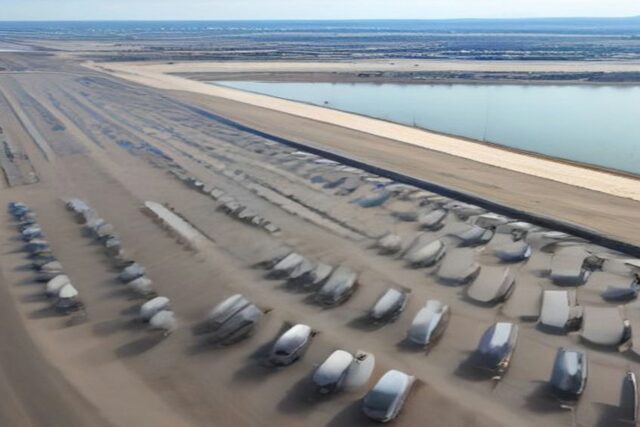 Vista aérea de un enorme patio con cientos de autos nuevos estacionados en filas junto a un cuerpo de agua en un día soleado