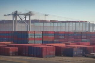 Patio portuario con largas pilas de contenedores de carga rojos y azules y una grúa de muelle al fondo bajo un cielo despejado