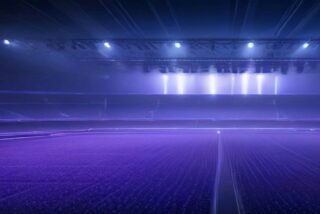 Estadio vacío de noche con filas de butacas iluminadas en tonos violeta y una estructura de reflectores que proyecta haces de luz blanca y azul, alusivo a los boletos de U2 para el 2 de marzo