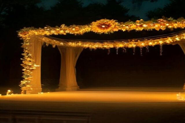 Escenario al aire libre con un arco de columnas cubierto de guirnaldas de cempasúchil y luces cálidas entre árboles de noche, alusivo al Día de Muertos INBAL 2026 en el Jardín Escénico
