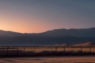 Cerca de postes y alambre en un llano seco del desierto al atardecer, con montañas y dunas al fondo, sin personas ni texto