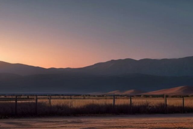 Cerca de postes y alambre en un llano seco del desierto al atardecer, con montañas y dunas al fondo, sin personas ni texto