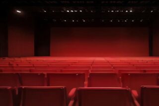 Auditorio vacío con hileras de butacas rojas, paredes oscuras y luces encendidas en el techo