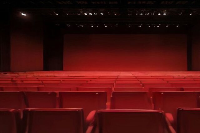 Auditorio vacío con hileras de butacas rojas, paredes oscuras y luces encendidas en el techo