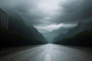 Avenida anegada y brillante por la lluvia entre hileras de árboles y edificios, con montañas cubiertas de nubes grises al fondo, sin personas ni texto