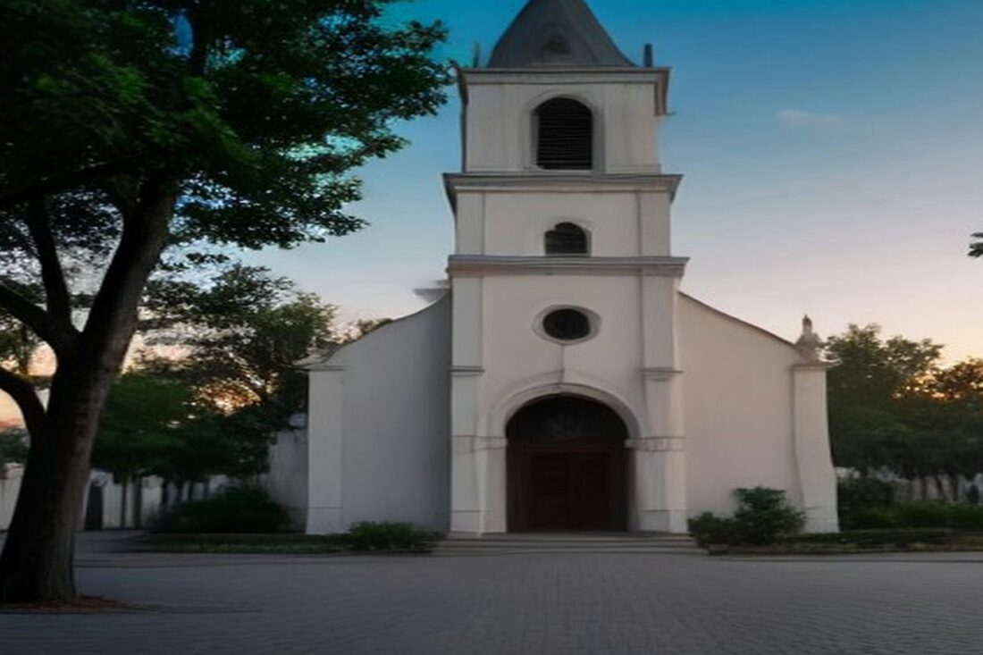 Iglesia colonial blanca con campanario frente a una plaza vacía y árboles, al atardecer