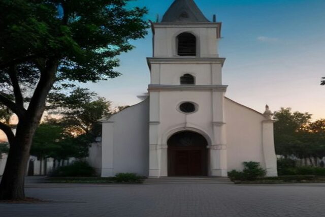 Iglesia colonial blanca con campanario frente a una plaza vacía y árboles, al atardecer