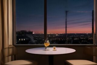 Habitación de hotel elegante con una mesa redonda, una copa y sillones frente a un gran ventanal con vista a una ciudad al anochecer