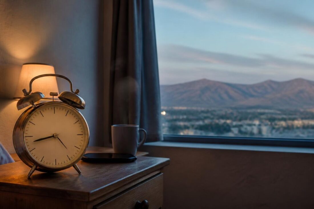 Reloj despertador analógico sobre un buró junto a una taza de café frente a una ventana con vista a una ciudad del norte de México al amanecer