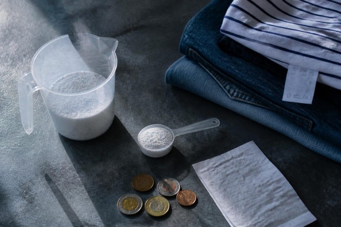 Taza medidora con detergente en polvo, cuchara dosificadora, monedas y ropa doblada con etiqueta de cuidado sobre una mesa
