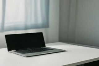 Computadora portátil abierta con pantalla oscura sobre un escritorio blanco junto a una ventana con cortina