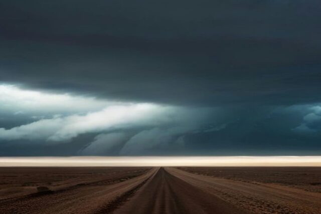 Nubes de tormenta oscuras y densas sobre una llanura desértica, con franjas de lluvia en el horizonte y un camino de tierra recto en primer plano