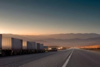 Carretera vacía al amanecer entre montañas con una fila de tractocamiones de caja blanca estacionados a un costado del camino, alusiva al paro de transportistas del 26 de octubre