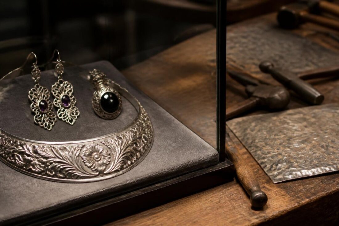 Collar de plata repujada con motivos florales y aretes calados con amatista en una vitrina de museo junto a cinceles de orfebre