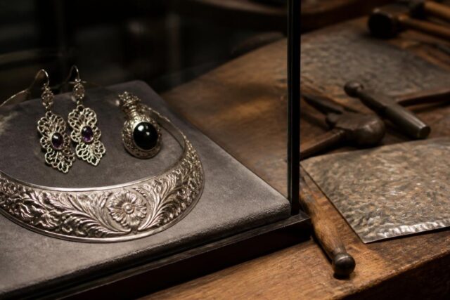 Collar de plata repujada con motivos florales y aretes calados con amatista en una vitrina de museo junto a cinceles de orfebre
