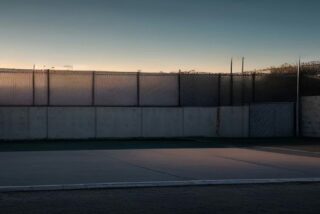 Explanada de concreto vacía junto a un muro y una malla ciclónica en un barrio al atardecer, con postes de luz y cielo en tonos azules