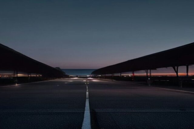 Vialidad vacía entre dos cubiertas largas de una terminal al anochecer con luces tenues en el horizonte, sin personas ni texto